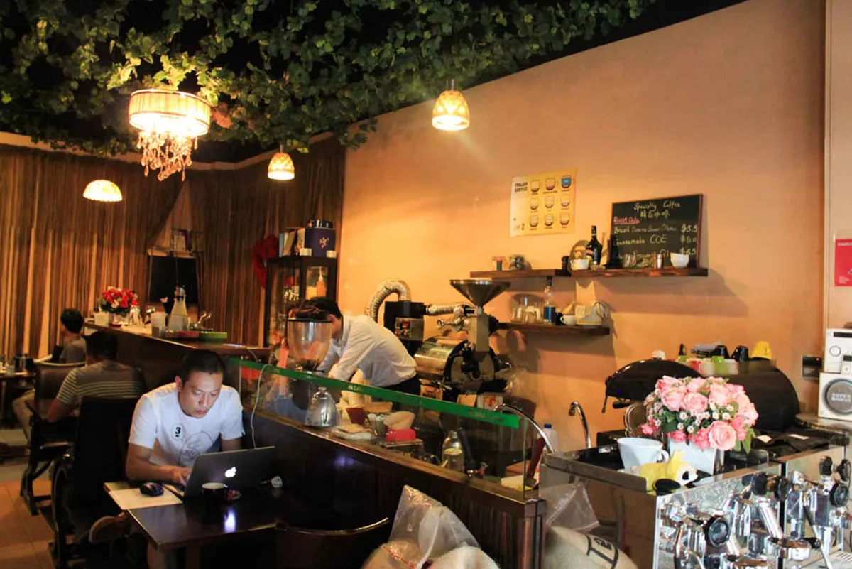 Cozy, dimly lit interior of Geisha Specialty Coffee Madrid featuring a professional coffee roaster, floral arrangements, and a customer working on a laptop, highlighting its reputation as one of the atmospheric study spots in Madrid.