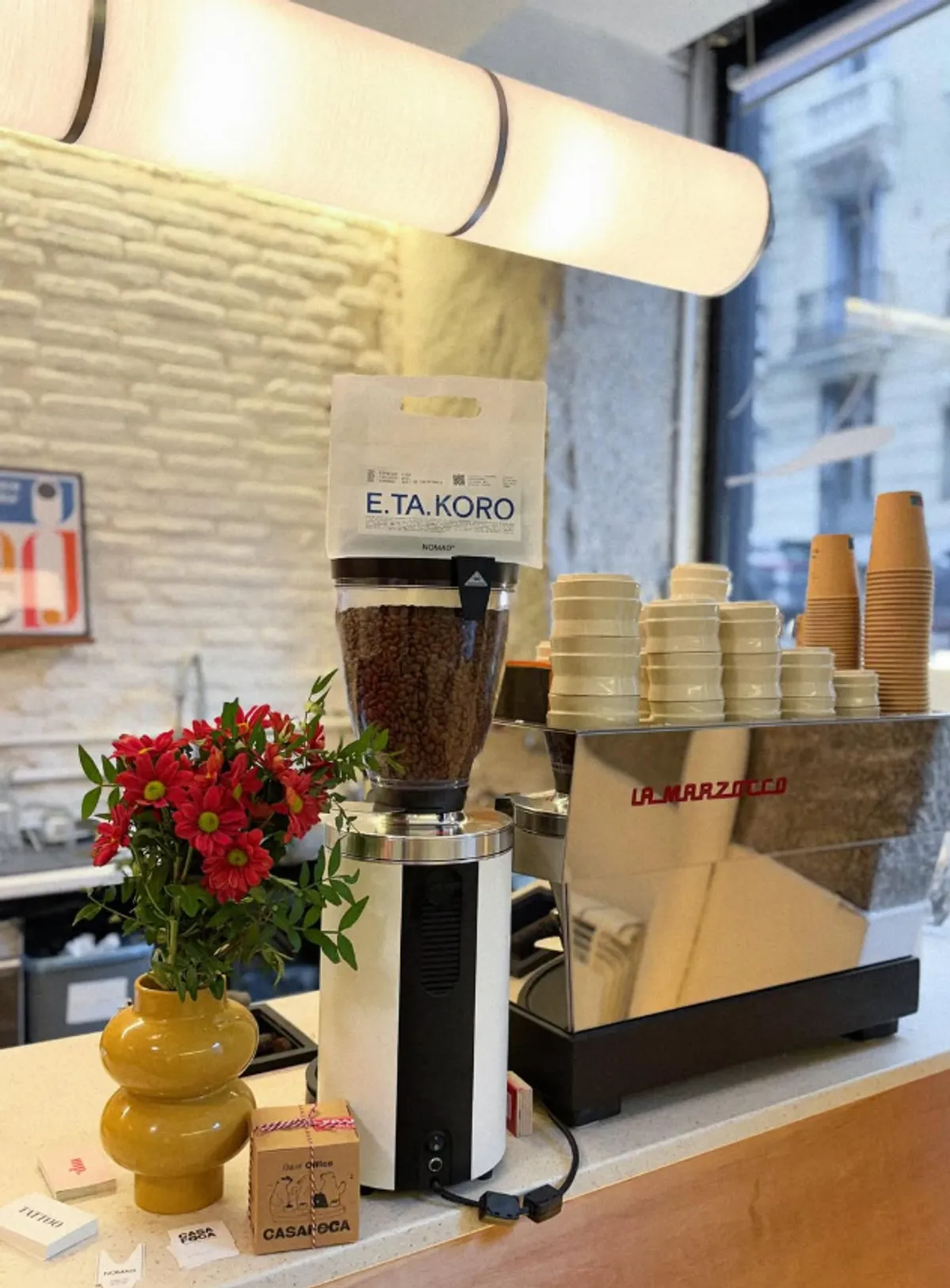 A professional coffee bar featuring a La Marzocco Linea espresso machine, a white coffee grinder with Nomad Coffee beans (E.TA.KORO), and fresh red flowers in a yellow vase.