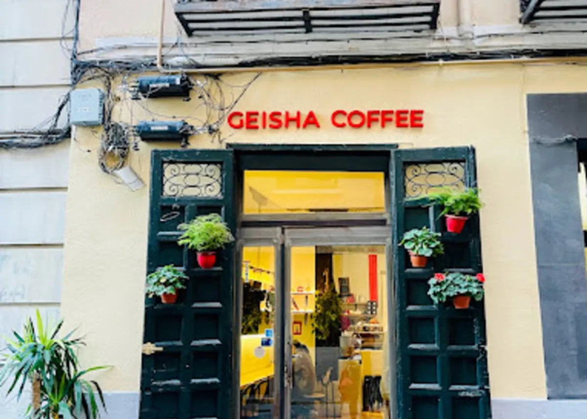 Exterior of Geisha Specialty Coffee, a minimalist cafe in Madrid with a pale yellow facade, bold red signage, and dark green wooden doors adorned with hanging plants—a quiet and inviting choice for local study spots in Madrid.