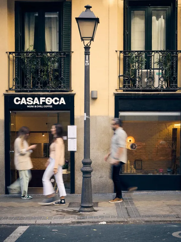 The minimalist black and white storefront of Casa Foca coffee shop in Madrid, featuring a traditional street lamp and a yellow building facade with wrought iron balconies