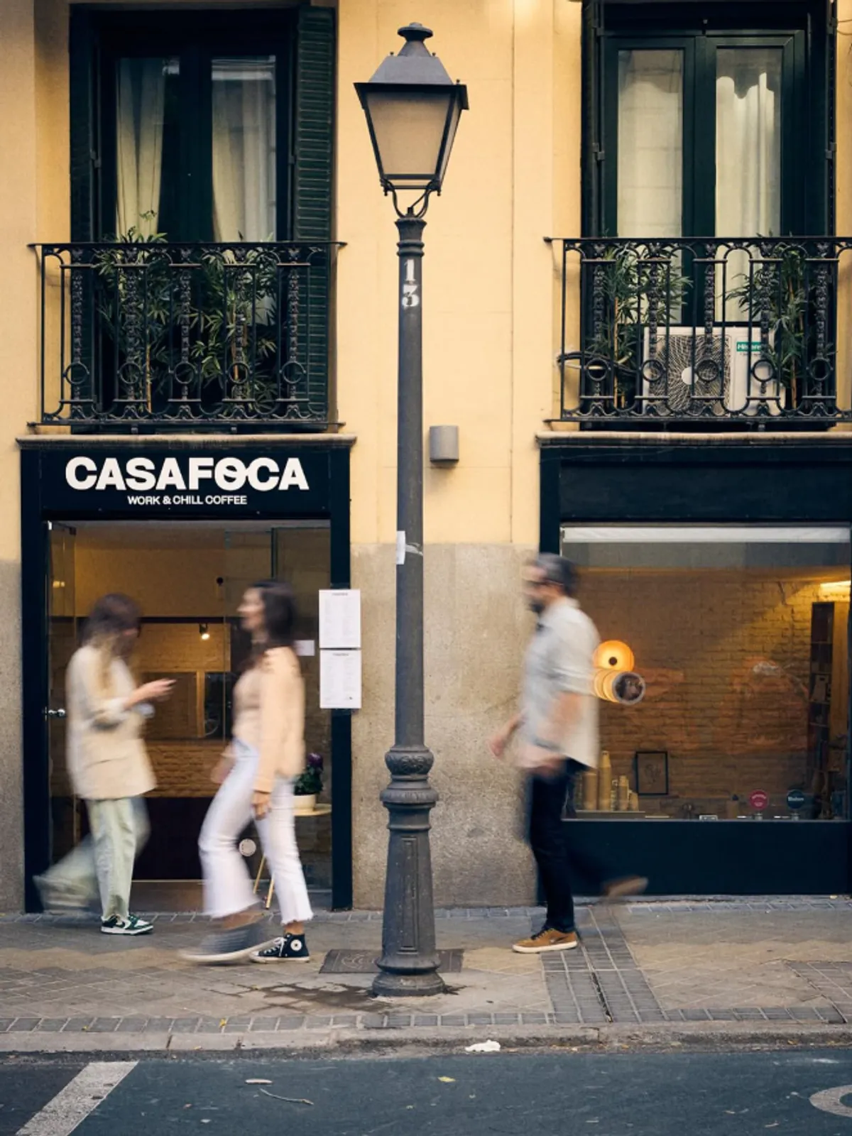 The minimalist black and white storefront of Casa Foca coffee shop in Madrid, featuring a traditional street lamp and a yellow building facade with wrought iron balconies