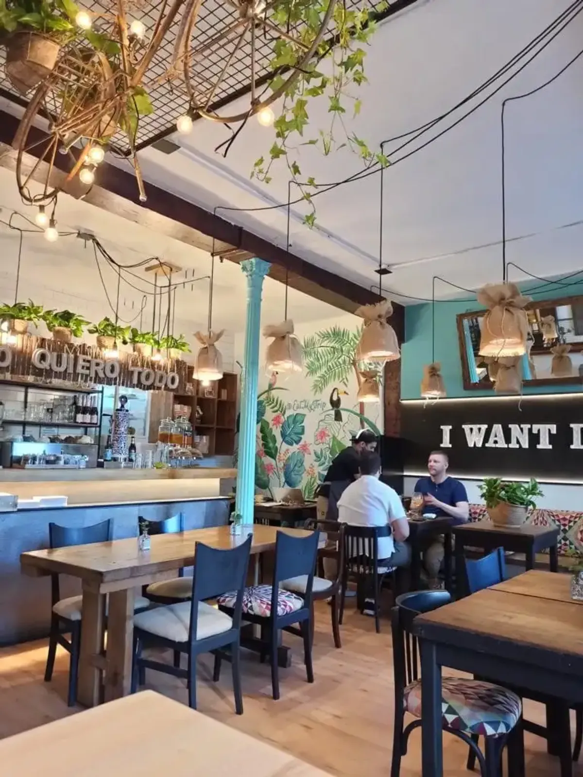 Spacious dining area at Eat My Trip cafe Madrid with a colorful tropical mural, "I WANT IT ALL" neon sign, and dedicated laptop-friendly seating, making it one of the top-rated creative study spots in Madrid.