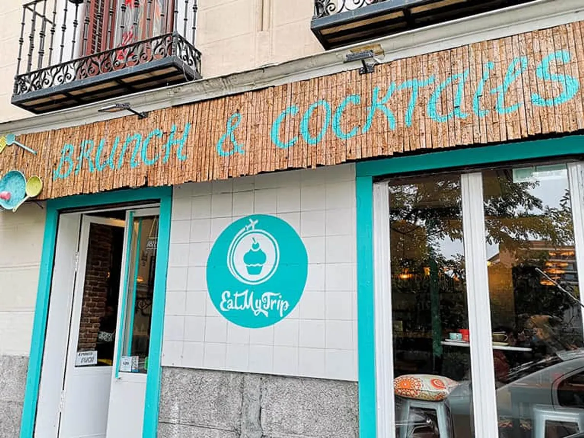 Vibrant exterior of Eat My Trip, a creative cafe in Madrid with bright turquoise trim and a "Brunch & Cocktails" bamboo sign, located in the heart of Malasaña—a popular destination for local study spots in Madrid.