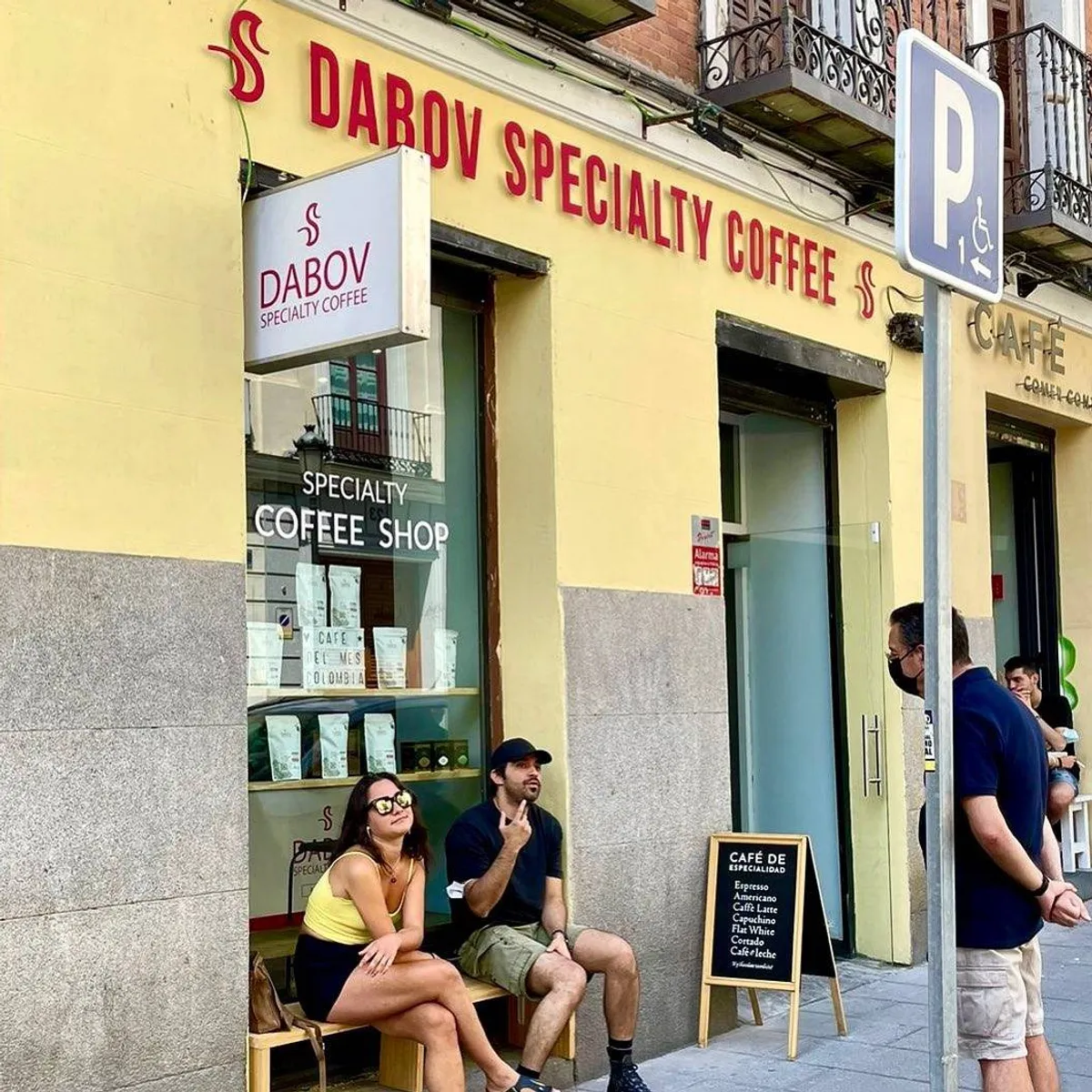 Customers sitting on benches outside Dabov Specialty Coffee, a minimalist cafe in Madrid with a bright yellow facade, large windows, and an outdoor menu—a top-rated choice for high-quality coffee and work-friendly study spots in Madrid.